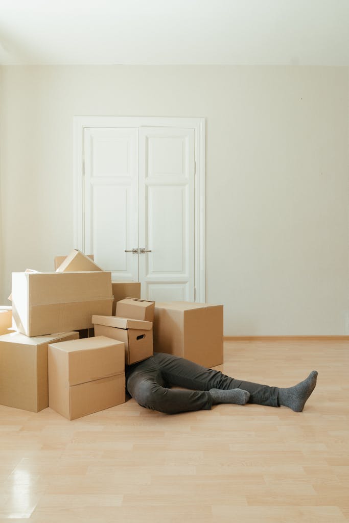 A person lying under a pile of moving boxes in a minimalistic room conveys tiredness from packing.