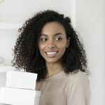 Young African American woman smiling while holding cardboard boxes indoors.